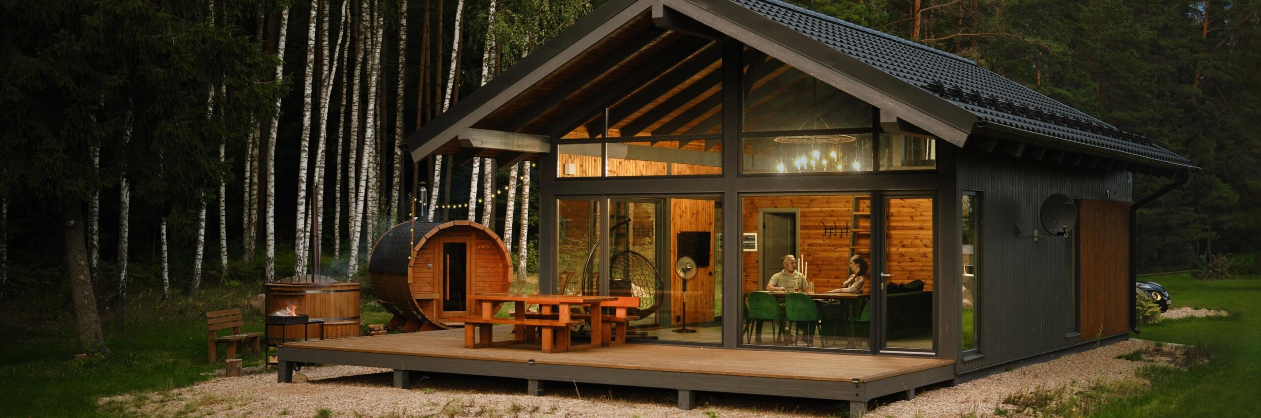 A modern cabin with a large glass windows and wooden deck with patio furniture at a forest clearing and a barrel-shaped wooden sauna