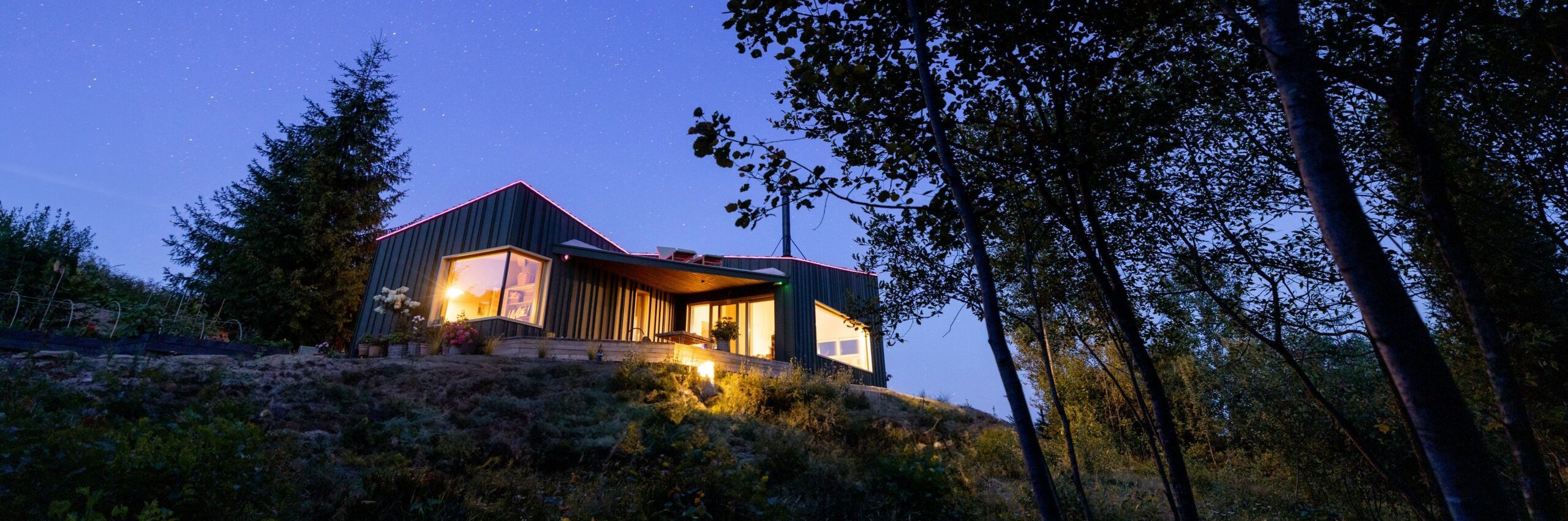 A cozy cabin illuminated warmly under a clear starry sky, framed by tree silhouettes. The view captures the serene atmosphere of this mountain retreat, perfect for stargazing and nature appreciation